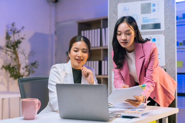 Obraz premium Two Asian businesswomen collaborating on financial reports while working late in a modern office, focused on their laptops and discussions
