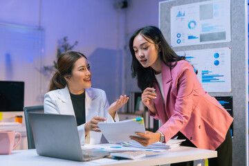 Two Asian businesswomen collaborating over a financial chart while working late at the office, analyzing data on a laptop