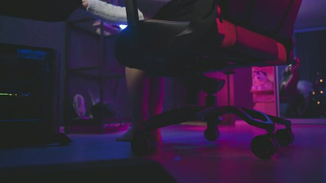 Low angle leg view of woman in fishnet pantyhose walking toward gaming chair and adjusting it closer to desk in neon lit studio, with glowing equipment and mirror reflecting seating area