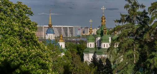 Scenic view of Vydubychi Monastery in Kyiv with golden domes and green rooftops, set against the modern skyline of the Dnipro’s left bank and lush spring trees.