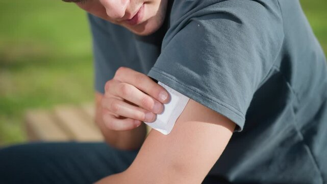 close up of white student seated outdoors pressing nicotine patch onto upper left arm while lifting shirt sleeve, with natural sunlight reflecting off skin and soft blur of green landscape in