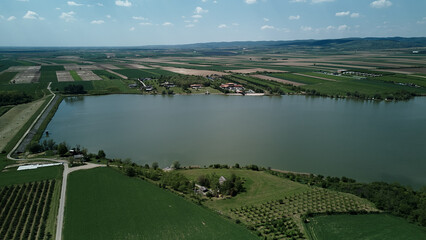 Drone view of Pavlovac Lake in Serbia surrounded by cultivated farmlands and villas. A scenic landscape of rural serenity, water, and agriculture under a clear spring sky.