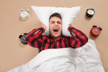 Young man suffering from loud noise of alarm clocks on beige background