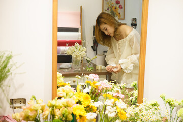 Asian woman arranging flowers in a cozy studio surrounded by vibrant blooms and crafting supplies...
