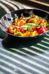 A colorful salad featuring vibrant tomatoes, cucumbers, and fresh herbs beautifully arranged in a black bowl. Sunlight casts playful shadows, enhancing the dish's visual appeal.