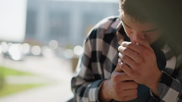 close up of smoker in black white plaid shirt holding cigarette between fingers cups hand around tip shields flame from breeze sparks ember against blurred cityscape background sunlit scene
