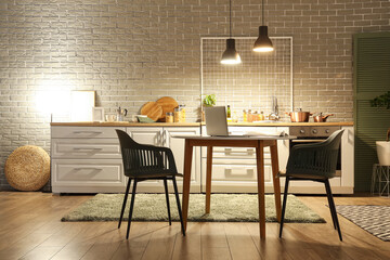 Interior of dark kitchen with counters, dining table and glowing lamps in evening