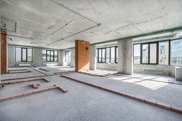 An expansive, empty commercial space shows signs of renovation, featuring concrete walls and a bright atmosphere created by large windows.
