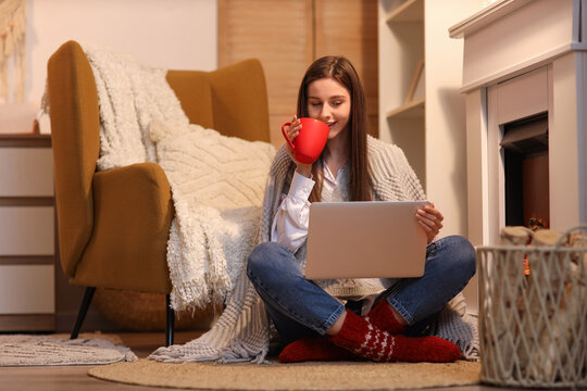 Young woman with tea and laptop sitting near fireplace at home in evening