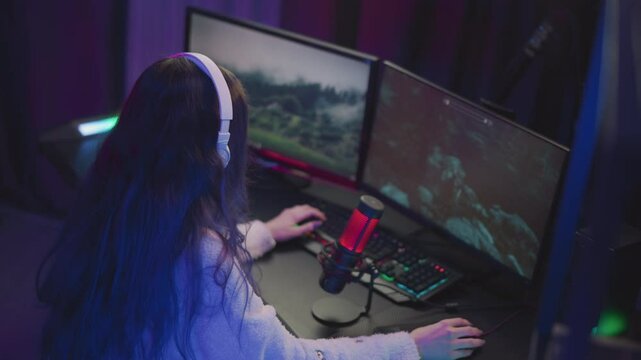 young woman with long wavy hair wearing white headset and fluffy sweater intensely playing game on dual monitors with red microphone and illuminated keyboard in dark colorful gaming room