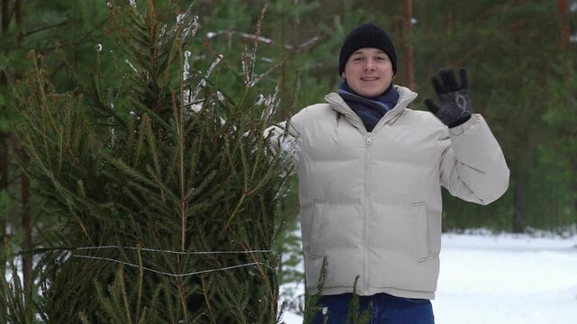 Man carrying christmas tree in snowy forest showing ok sign. A guy waves his hand affably next to a fir tree in the forest