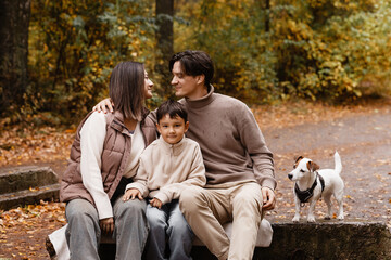 Fototapeta premium Front view of young parents and son sitting on a park bench with small dog, autumn forest background. Concept of family, active lifestyle, pet care, family health, mortgage and family security.