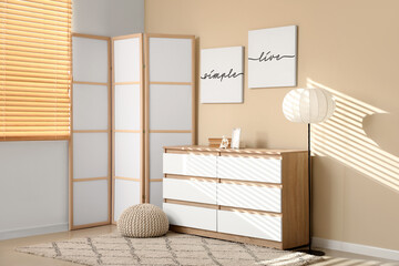 Interior of light living room with folding screen and chest of drawers near beige wall