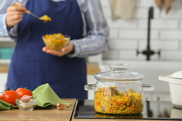 Cooking pot with pilaf on stove against woman in kitchen, closeup