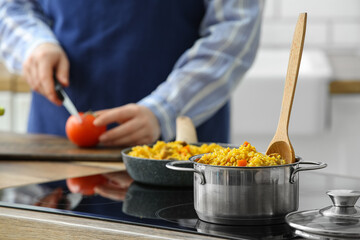 Cooking pot with pilaf on stove against woman cutting tomato in kitchen, closeup