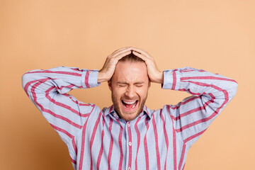 Young man with hands on head showing frustration against beige background, wearing striped shirt, expressing emotions authentically