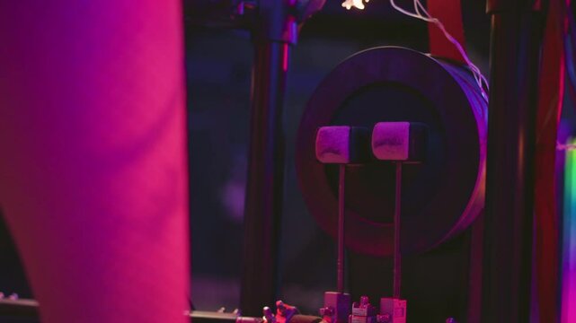 Close up of leg in fishnet pantyhose pressing twin beaters against kick drum pedal in studio setting with pink and purple neon lighting and surrounding wires during music practice session