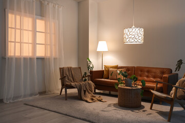 Interior of living room with brown sofa, armchairs and glowing lamps in evening