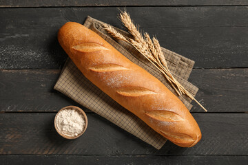 Fresh baguette with wheat and bowl of flour on black wooden background