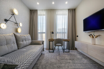 A cozy living room features a gray patterned sofa, a sleek desk with a chair, and large windows allowing natural light to fill the space. Decorative lighting enhances the modern ambiance.