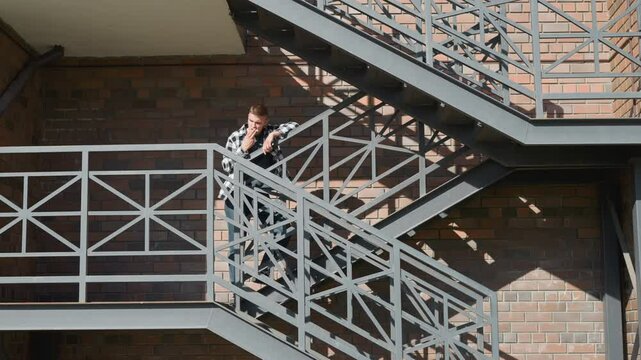 Young man in black and white checkered shirt stands on metal balcony staircase smoking cigarette with relaxed posture against brick wall as diagonal sunlight casts shadows on patterned iron railing
