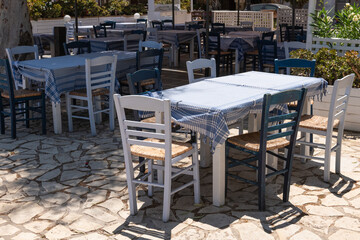 Traditional outdoor Greek taverna setup with blue checkered tablecloths and wooden chairs under sunlight.