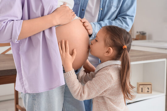 Cute little girl kissing her pregnant mother's belly and father in kitchen