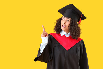 Surprised African-American female graduate pointing at something on yellow background