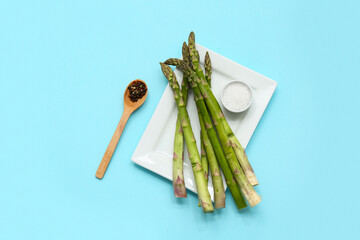 Plate with fresh green asparagus, peppercorns and bowl of salt on blue background