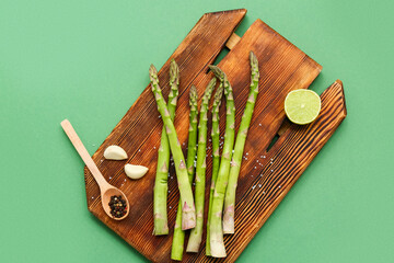 Wooden board with fresh green asparagus, lime, peppercorns and garlic on color background