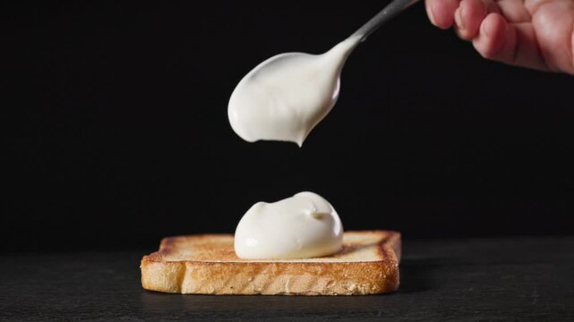 A cook put mayonnaise on fresh toasted bread slice on black wooden board for making club sandwich, food closeup. Process of making healthy meal