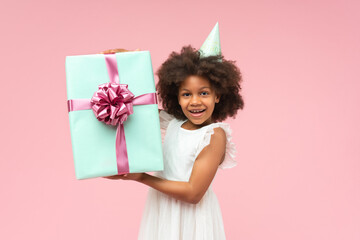 Adorable African American girl wearing white ruffle dress and birthday hat posing over pastel pink background with decorated big present box in her hands with excited face expression