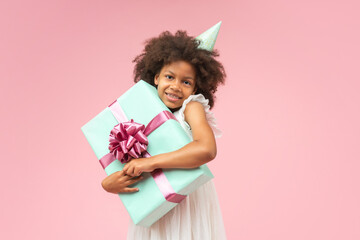 Portrait of adorable African American birthday girl posing in studio over pastel pink background hugging big present box and looking at the camera with cute smile