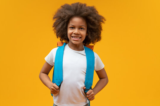 Studio shot of a cute cheerful African American schoolgirl posing over yellow background smiling to the camera and holding straps of her backpack