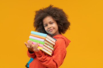 Studio shot of lovely and sweet African American little girl student holding pile of colored books in hands and looking to the camera isolated over yellow background