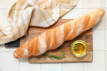 Wooden board with fresh baguette, rosemary and glass bowl of oil on white tile background