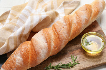 Wooden board with fresh baguette, rosemary and glass bowl of oil on white tile background, closeup