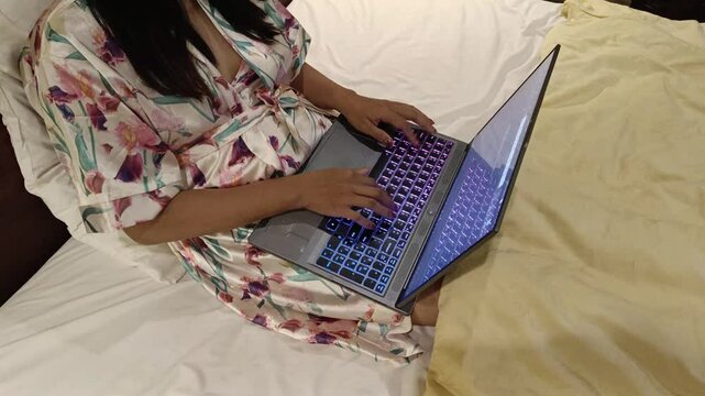 A woman is working on her laptop in bed, wearing a robe with a floral pattern and a keyboard with colorful illumination. It's a cozy and casual scene.