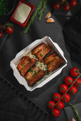 grilled pork belly slices with herbs and onions in takeaway box on dark background with tomatoes and spices