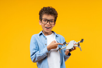 Studio shot of adorable African American schoolboy pointing with index finger at the drone in his hand with excited open mouth face expression, isolated over orange yellow background