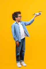 Full length vertical shot of a kid student posing in studio holding generic design drone in his hand, isolated over bright colored orange yellow background