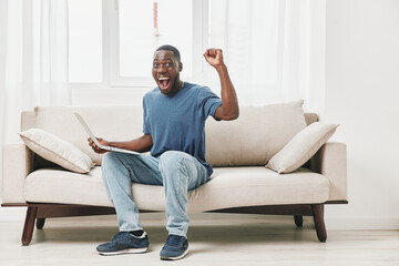 Excited young African American man celebrating success while sitting on a cozy sofa with a laptop...