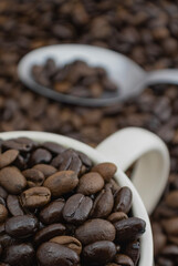 Up close macro of espresso beans in a cream colored cup, with a spoon in the background. Edge to edge coffee beans make up the background as well. Shallow depth of field.