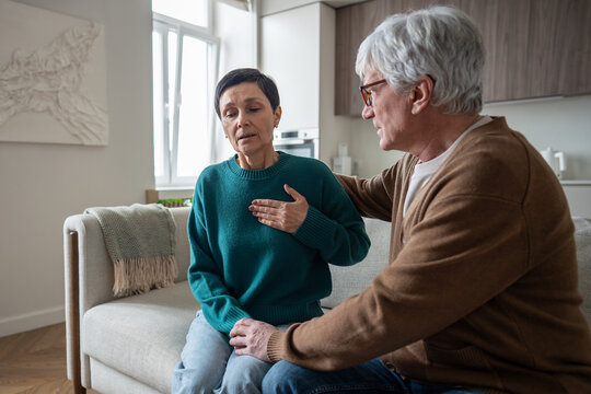 Struggling worried senior woman clutches chest, breathes heavily while man gently supports on home sofa. Asthma labored breathing, chest pain and irregular heartbeat, emotional panic symptoms