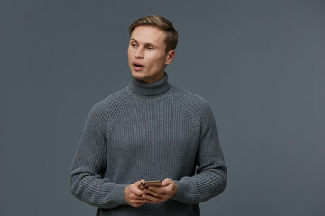 Man with short brown hair wearing gray turtleneck sweater holding smartphone, standing against plain gray background. Casual indoor portrait with focused expression, suitable for technology
