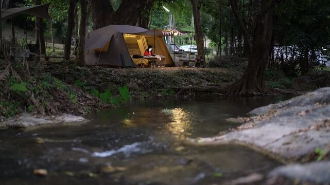 nature camping and camper relaxing in front of tent with warm white lamp on evening twilight and water stream at garden forest lan sang waterfall park campground on holiday vacation with stream sound