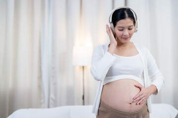Asian pregnant woman wearing headphones smiling softly eyes closed gently cradling belly standing...