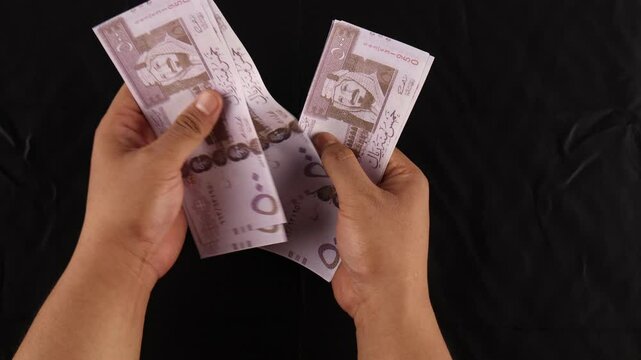 close up of a man counting 500 riyal banknotes with a black background.
