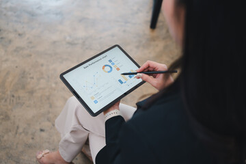 Businesswoman analyzing sales performance chart on digital tablet, using stylus pen, sitting on...
