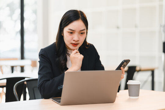 Asian businesswoman black suit sitting at desk looking at laptop with worried expression holding smartphone coffee cup on table modern office background stress at work